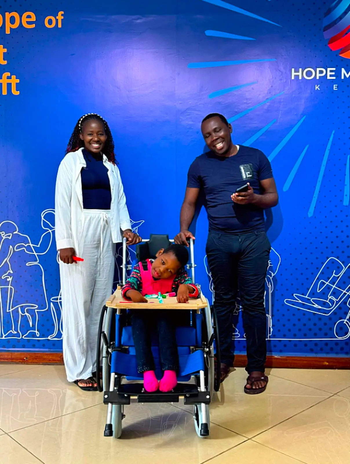 A young child smiling in a new specialized wheelchair, with two Nyasuba Golden Pillars volunteers